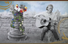 Stephen Stills sits next to a tower covered in flowers. In his lap is an acoustic guitar. This image is a screen grab from the Youtube Video
